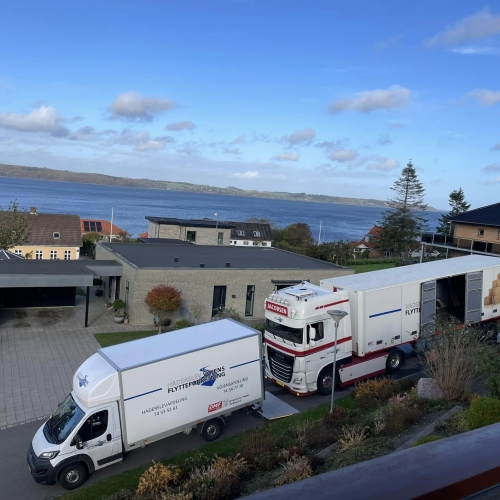 To flyttebiler, en stor lastbil og en flyttebil fra Haderslev og Vojens flytteforretning, parkeret foran en moderne bolig med udsigt over en fjord. Bilerne holder i en indkørsel omgivet af velplejet have og grønne buske.