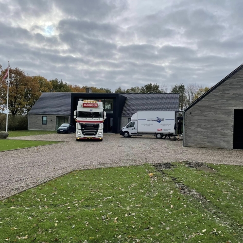 En stor flyttelastbil og en mindre flyttebil fra Haderslev og Vojens flytteforretning parkeret foran et moderne, gråt stenhus med sort tag i et landligt område.