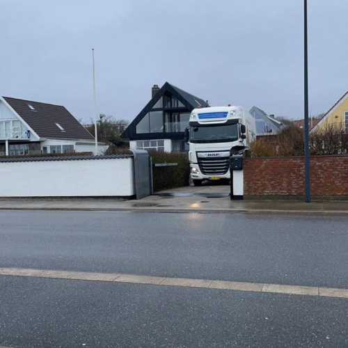 En stor flyttelastbil fra Haderslev og Vojens flytteforretning holder parkeret foran indkørslen til et moderne sort-hvidt hus i et boligområde på en grå dag..