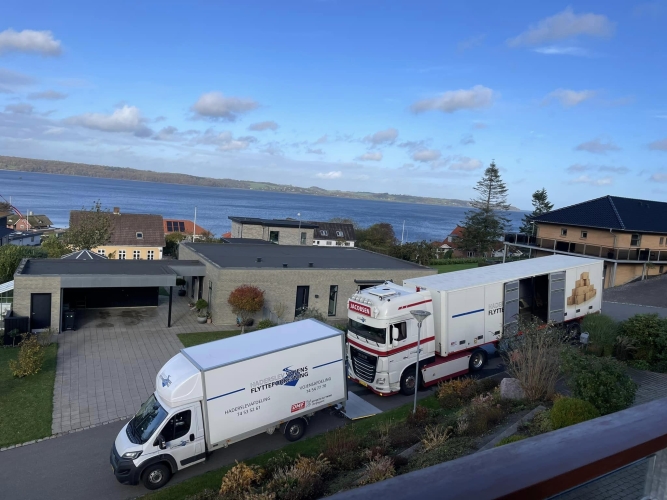 To flyttebiler, en stor lastbil og en flyttebil fra Haderslev og Vojens flytteforretning, parkeret foran en moderne bolig med udsigt over en fjord. Bilerne holder i en indkørsel omgivet af velplejet have og grønne buske.