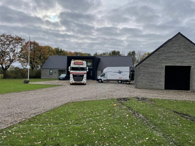 En stor flyttelastbil og en mindre flyttebil fra Haderslev og Vojens flytteforretning parkeret foran et moderne, gråt stenhus med sort tag i et landligt område.