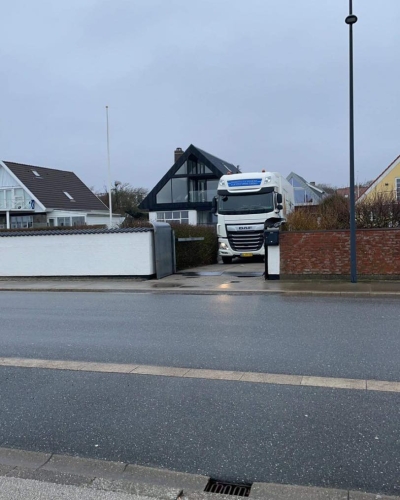 En stor flyttelastbil fra Haderslev og Vojens flytteforretning holder parkeret foran indkørslen til et moderne sort-hvidt hus i et boligområde på en grå dag..